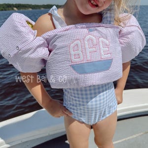May include: A light purple and white seersucker life vest with the initials "BFE" embroidered on the front, above a light blue boat design. The vest is worn over a light blue gingham swimsuit. The text "West Bai & Co" is visible.