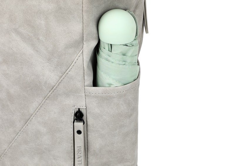 K&ouml;nnte beinhalten: Ein grauer Rucksack mit einer Rei&szlig;verschlusstasche an der Seite. Ein hellgr&uuml;ner Regenschirm ist in die Tasche gesteckt. Der Rei&szlig;verschluss hat den Schriftzug "BRAT" aufgedruckt.
