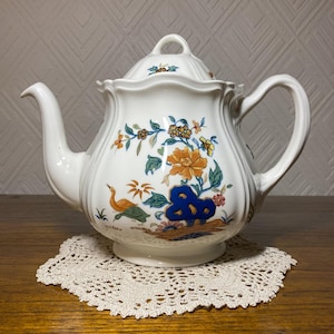 Puede incluir: Tetera blanca con un diseño floral en tonos azules, amarillos, naranjas y verdes. La tetera tiene una tapa y un asa.