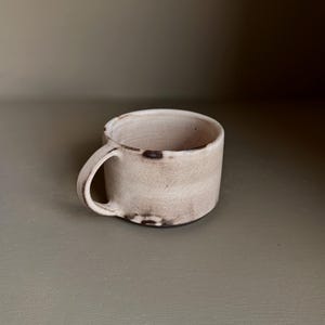 May include: A small, low-profile ceramic mug with a light beige, speckled finish. The mug features a single curved handle on the side and dark brown, irregular markings along the base and rim, giving it a textured, hand-crafted appearance.