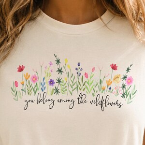 以下が含まれることがあります： クリーム色のTシャツ。カラフルな花の模様と「you belong among the wildflowers」の文字がプリントされています。デザインにはピンク、赤、黄色、紫の色合いの花が含まれています。Tシャツはウエストで結ばれています。