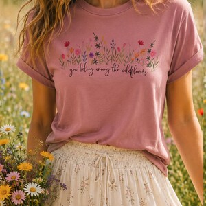 以下が含まれることがあります： ダスティーローズのTシャツ。花柄のデザインと「you belong among the wildflowers」の文字入り。袖はロールアップされています。モデルは花柄のスカートを着用しています。