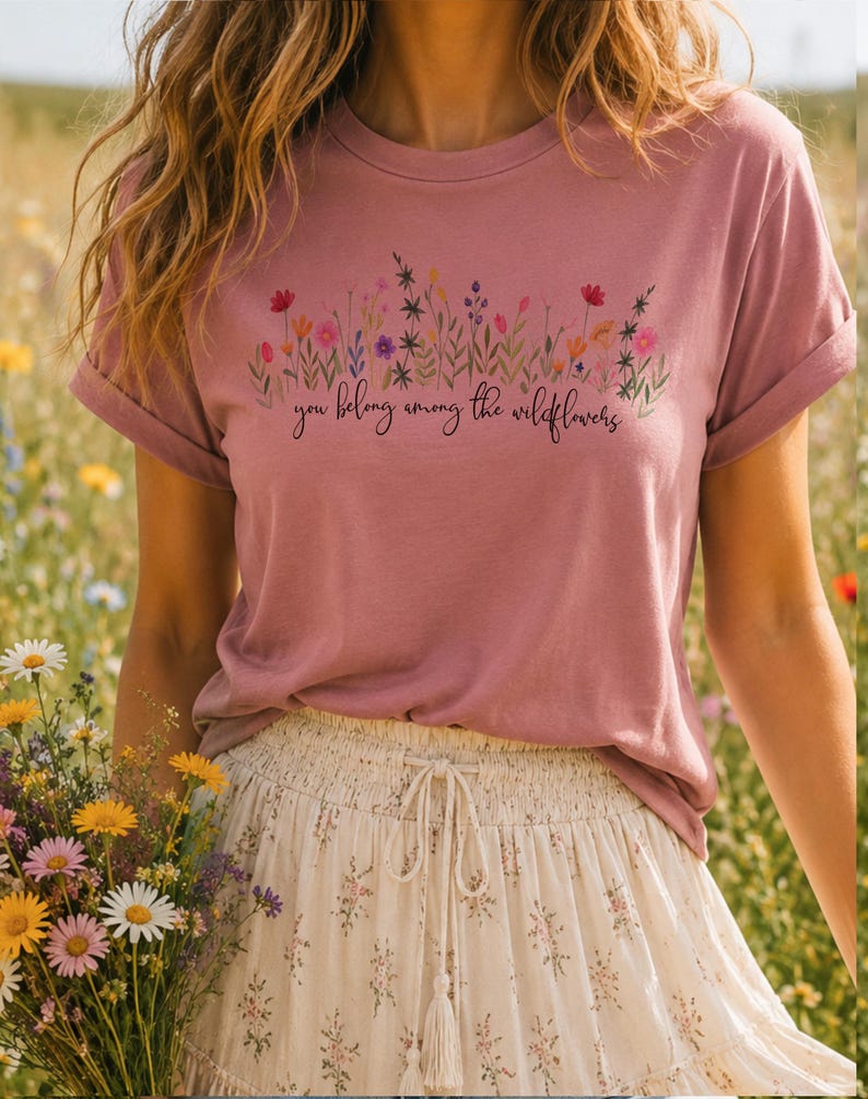 以下が含まれることがあります： ダスティーローズのTシャツ。花柄のデザインと「you belong among the wildflowers」の文字入り。袖はロールアップされています。モデルは花柄のスカートを着用しています。
