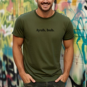 May include: A man wearing a green t-shirt with the text "Ayuh, bub." printed on the front. He is standing in front of a colorful graffiti wall.