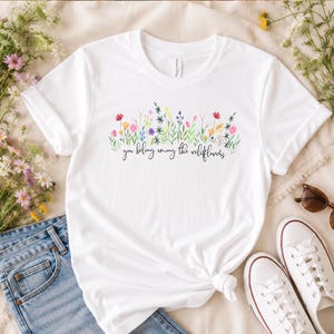 以下が含まれることがあります： 水彩画の花柄と「you belong among the wildflowers」の文字が入った白いTシャツ。青いジーンズと白いスニーカーと合わせて、カジュアルな普段着スタイルを提案しています。リラックスした雰囲気で、毎日の着こなしに最適です。