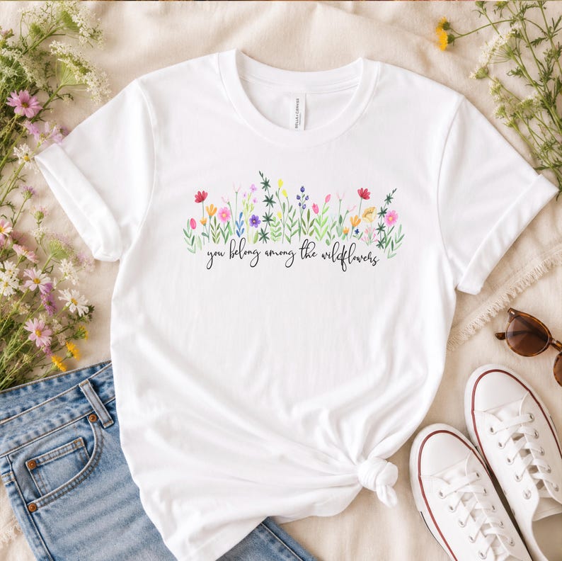以下が含まれることがあります： 水彩画の花柄と「you belong among the wildflowers」の文字が入った白いTシャツ。青いジーンズと白いスニーカーと合わせて、カジュアルな普段着スタイルを提案しています。リラックスした雰囲気で、毎日の着こなしに最適です。