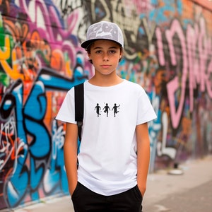May include: A young person wearing a white t-shirt with a black graphic of three stick figures playing football. The person is also wearing a grey mesh baseball cap and black trousers. The person is standing in front of a graffiti wall.