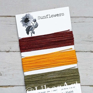 May include: A card with three bundles of thread in rust, yellow-orange, and khaki colors. The card has the word "sunflowers" and a sunflower illustration. The text "Rust - Yellow-Orange - Khaki" is also visible.
