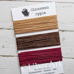 May include: A card with three colors of thread: butterscotch, walnut, and red. The card is labeled "Cinnamon Apple" with an apple and cinnamon stick illustration. The text "UberArt" and "Five yards of each color" are also visible.