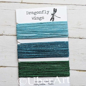 May include: A card labeled "Dragonfly Wings" displays three colors of embroidery floss: turquoise, teal, and emerald green. Each color is neatly wound and separated on the card. The card also features a dragonfly illustration.