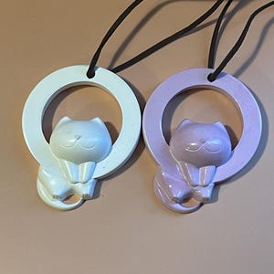 May include: Two ceramic cat ornaments hanging from black cords. The cat on the left is white and the cat on the right is light purple. Both cats are holding onto a circular ring.