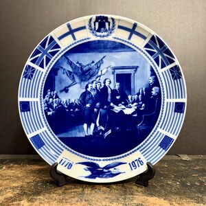 May include: A round ceramic commemorative plate featuring a blue and white transferware scene of the signing of the Declaration of Independence. The border displays various historical flags and the Liberty Bell. The dates 1776 and 1976 are printed.