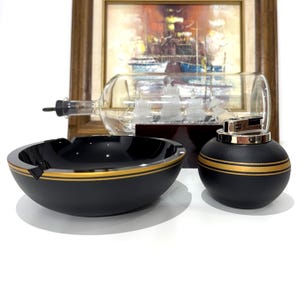 Pode incluir: Um cinzeiro e um isqueiro pretos com detalhes dourados. O cinzeiro é redondo com uma área rebaixada para cigarros. O isqueiro esférico tem uma faixa dourada. Um navio de vidro numa garrafa está no fundo.