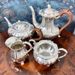 Op de afbeelding: Een verzameling antieke zilveren theeserviesartikelen. De set bevat een theepot met een houten handvat, een melkkan, een suikerpot en een kleinere theepot. De stukken hebben sierlijke details en zijn gerangschikt op een houten oppervlak.