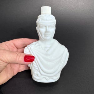 May include: A white, vintage perfume bottle shaped like a bust of a Roman figure. The bottle has a white plastic cap and is held by a hand with red nail polish. The bust is detailed with facial features and draped clothing.