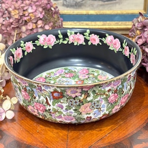 Può includere: Una ciotola decorativa in ceramica con interno nero e design floreale. Il bordo è adornato da rose rosa e l'esterno presenta un motivo dettagliato di fiori e uccelli. La ciotola ha un bordo dorato.