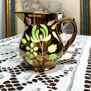 May include: A small, antique, gold-toned jug with a floral design in shades of green, pink, and yellow. The jug has a curved handle and a spout. The background includes a white lace tablecloth and a framed picture.