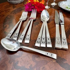 Pode incluir: Um conjunto de talheres prateados incluindo garfos, colheres e facas com um padrão floral decorativo nos cabos. Os utensílios apresentam estrias verticais ao longo das hastes e um acabamento polido sobre uma superfície de madeira escura.