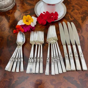 Può includere: Un set di posate color argento composto da quattro cucchiai, quattro forchette e quattro coltelli, ciascuno con un manico decorativo in rilievo. Le posate sono disposte su una superficie in legno accanto a una tazza, piattino e fiori.