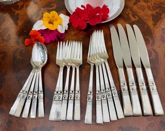 Peut inclure: Un ensemble de couverts argentés comprenant quatre cuillères, quatre fourchettes et quatre couteaux, chacun doté d'un manche décoratif en relief. Les couverts sont disposés sur une surface en bois avec une tasse, une soucoupe et des fleurs.