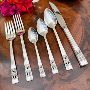 Pode incluir: Um conjunto de cinco talheres de aço inoxidável, incluindo um garfo de mesa, um garfo de salada, uma colher de chá, uma colher de sopa e uma faca. Cada peça apresenta um cabo com design canelado e um motivo floral na base.
