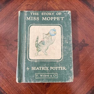 May include: A vintage copy of "The Story of Miss Moppet" by Beatrix Potter. The book has a green cover with the title in white and a framed illustration of a cat. The cat is holding a blue object. The book is published by F. Warne & Co.