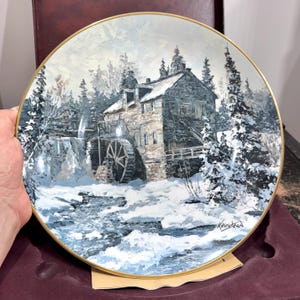 May include: Decorative plate featuring a winter scene with a stone mill, snow-covered trees, and a flowing river. The plate has a gold rim and is signed by Keirstead. The artwork depicts a serene, snowy landscape.