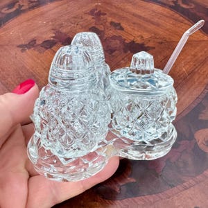 Peut inclure: Un ensemble à condiments en verre transparent comprenant deux salières et un pot couvert avec une petite cuillère, le tout posé sur un plateau en verre assorti. Chaque pièce présente un motif en losange texturé et le pot inclut une cuillère.