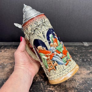 May include: A ceramic beer stein with a detailed relief design. The stein features a silver metal lid and handle. The body is decorated with a scene of people in colorful clothing. The stein has a beige and brown color scheme.