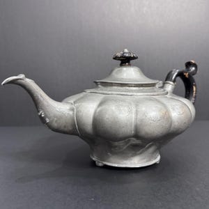 May include: Antique pewter teapot with a bulbous body and a black handle. The teapot has a decorative lid with a finial and a curved spout. The teapot is a vintage item, likely from the early 20th century.