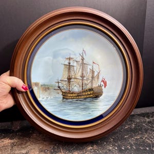 May include: A round decorative plate featuring a detailed painting of a tall ship sailing on the ocean. The plate is framed with a dark brown wooden frame, a gold inner ring, and a blue border. The ship has multiple sails and red flags.