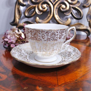 May include: White teacup and saucer set with gold floral detailing. The teacup has a curved handle and sits on a matching saucer. The set is placed on a wooden surface, with a decorative gold accent in the background. A vintage tea set.