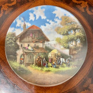 Può includere: Un piatto decorativo in ceramica con una pittura dettagliata di una scena rurale. Raffigura una fattoria, persone, cavalli e balle di fieno sotto un cielo blu con nuvole. Il piatto ha una finitura lucida e un bordo marrone.