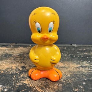 Op de afbeelding: Een vintage gele Tweety Bird-beeldje met oranje voeten en snavel. Het stripfiguur heeft grote blauwe en witte ogen en staat rechtop. Het beeldje is gemaakt van een glanzend materiaal en is een verzamelobject.