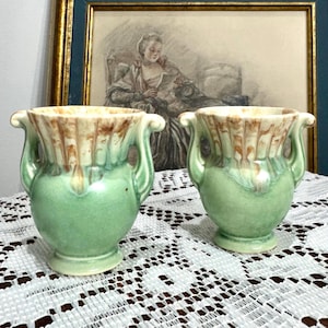 Peut inclure: Deux petits vases en céramique assortis. Chaque vase a une base verte, un bord marron et crème, et deux anses incurvées. Les vases sont présentés sur une nappe en dentelle blanche, avec un tableau encadré en arrière-plan.