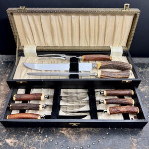 Può includere: Un set da intaglio in una scatola nera con rivestimento in raso color crema. Il set include una forchetta a due rebbi, un coltello grande, un acciaino e sei coltelli piccoli, tutti con manici marroni dalla texture ruvida.