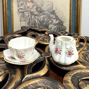 May include: A vintage porcelain tea set comprising a teacup, saucer, and small teapot. The white items are decorated with pink and blue floral patterns and gold edging. The teacup and teapot have a fluted design.