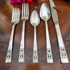 Pode incluir: Um conjunto de cinco talheres de aço inoxidável composto por um garfo pequeno, um garfo grande, uma colher de chá, uma colher de sopa e uma faca. Cada peça apresenta um cabo com nervuras verticais e um motivo floral na base.