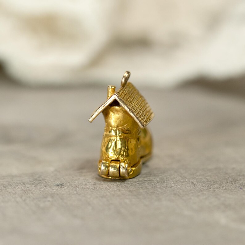 May include: A gold-colored charm in the shape of a boot with a house on top. The boot has intricate details, and the house features a textured roof. The charm is likely made of gold-toned metal and is designed to be a pendant.