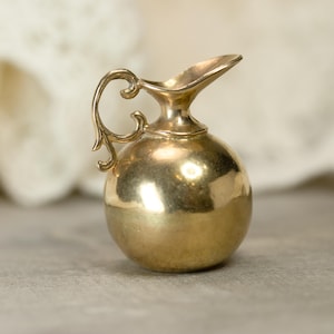 Puede incluir: Una pequeña jarra dorada con cuerpo redondeado y asa curvada. La jarra tiene un pico para verter y probablemente está hecha de metal. El asa tiene un diseño decorativo y ornamentado. El estilo general sugiere una estética vintage o antigua.
