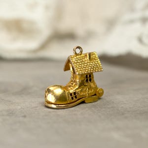 May include: A gold-colored charm in the shape of a boot with a house on top. The boot has a rounded toe and small windows. The house has a textured roof and a chimney. The charm has a loop at the top for attaching to a bracelet or necklace.