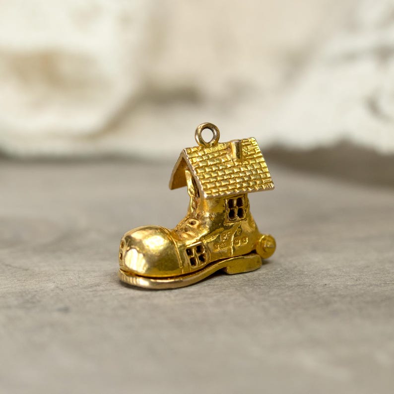 May include: A gold-colored charm in the shape of a boot with a house on top. The boot has a rounded toe and small windows. The house has a textured roof and a chimney. The charm has a loop at the top for attaching to a bracelet or necklace.