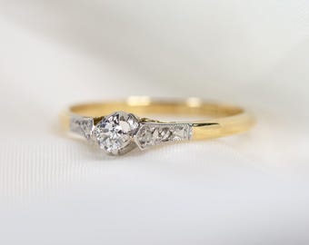 Puede incluir: Un delicado anillo de compromiso de oro y plata con un solo diamante redondo en el centro. La banda está adornada con diamantes más pequeños.
