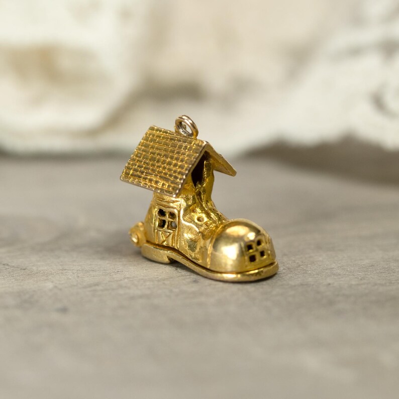 May include: A gold-colored charm in the shape of a boot with a house on top. The boot has a rounded toe and small windows. The house has a textured roof. The charm has a loop at the top for attaching to a necklace or bracelet.
