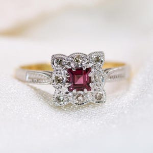 May include: A vintage ring featuring a square, deep red gemstone, encircled by smaller, clear stones. The silver band has detailed patterns, with a gold interior. A classic piece of jewellery.