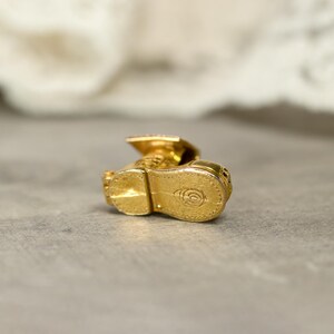 May include: A small, gold-colored charm in the shape of a graduation cap and gown. The charm is highly detailed, with visible textures and a shiny finish. The sole of the shoe has a circular design. The charm is likely made of metal.