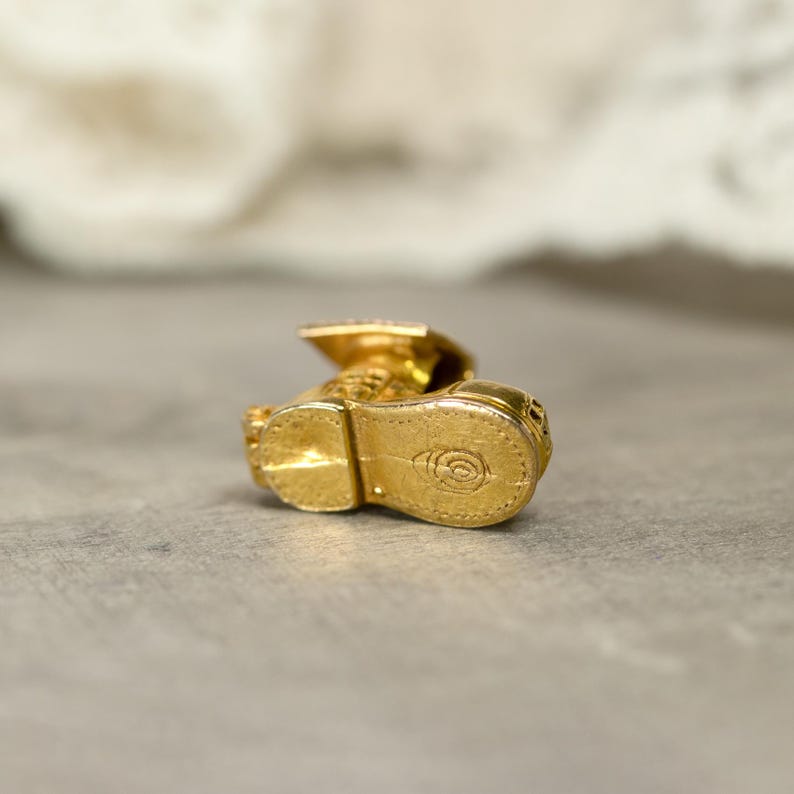 May include: A small, gold-colored charm in the shape of a graduation cap and gown. The charm is highly detailed, with visible textures and a shiny finish. The sole of the shoe has a circular design. The charm is likely made of metal.