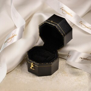 May include: An open, black, octagonal jewelry box with gold trim and a gold clasp. The interior is lined with black velvet. A white ribbon with gold text is draped around the box.