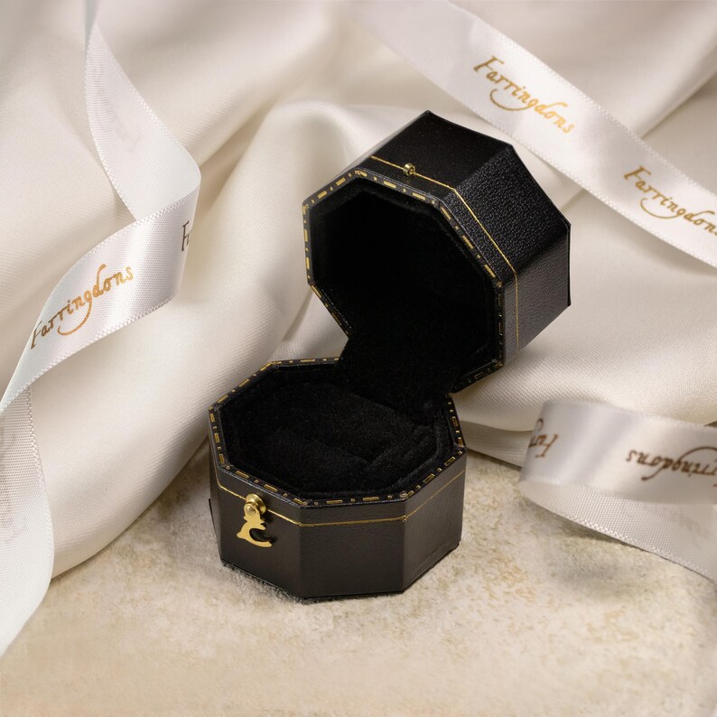 May include: An open, black, octagonal jewelry box with gold trim and a gold clasp. The interior is lined with black velvet. A white ribbon with gold text is draped around the box.