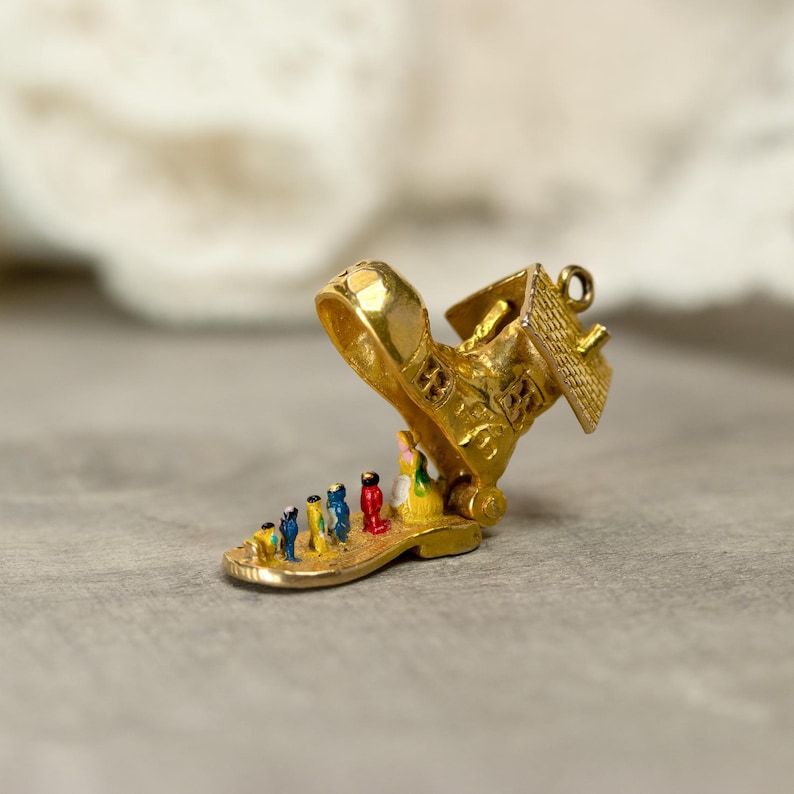 May include: A gold-colored charm in the shape of a shoe with a house on top. The shoe's sole features a line of tiny, colorful figures. The house has a detailed roof and windows. A small loop is attached for hanging.